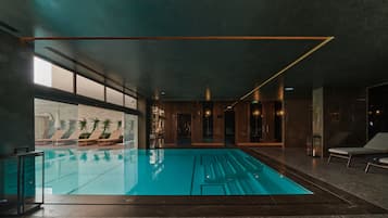 Una piscina interior moderna con paredes de vidrio transparente, sillas de descanso y vistas a un área exterior con plantas y sombrillas.