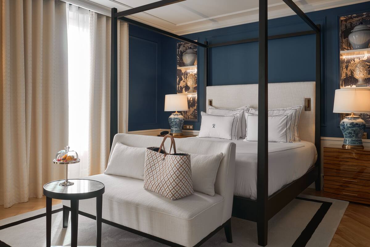 A hotel room with a four-poster bed, a round side table with a lamp, a sofa, and a decorative tray with fruit.