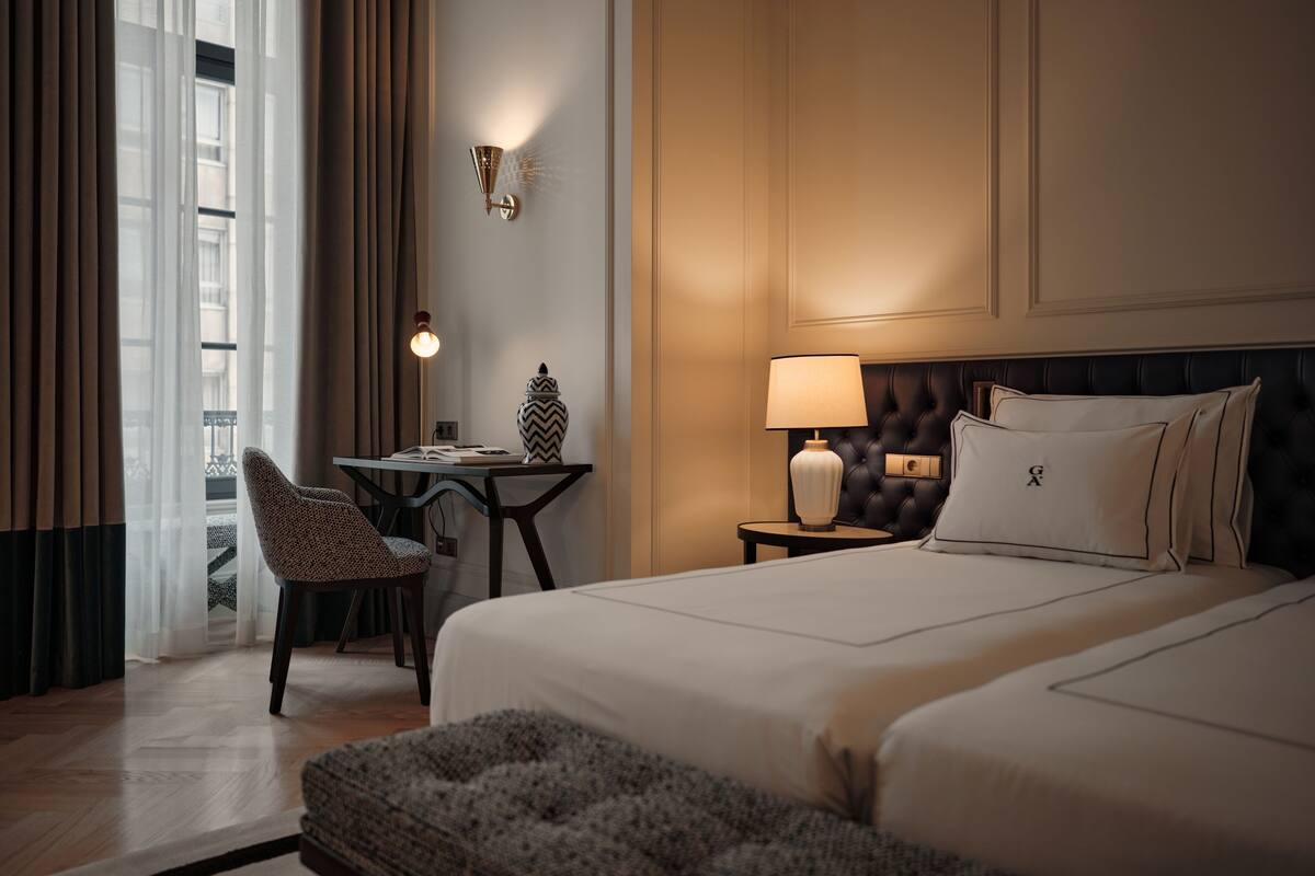 A hotel room with a large bed, a desk with a chair, a lamp, and a view of the outside through a window with curtains.