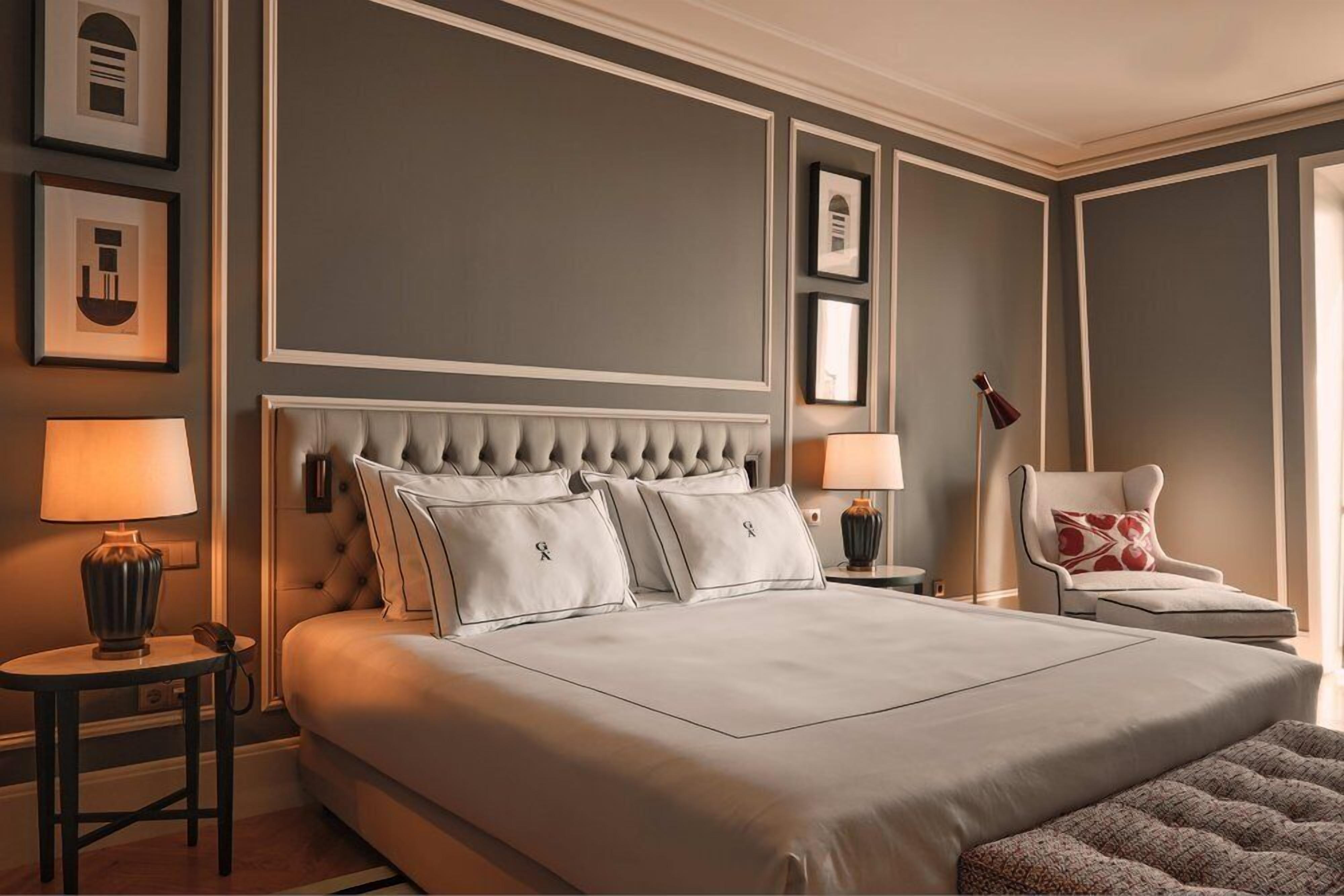 A large bed with a tufted headboard, two bedside tables with lamps, a chair with a cushion, and framed pictures on the wall.