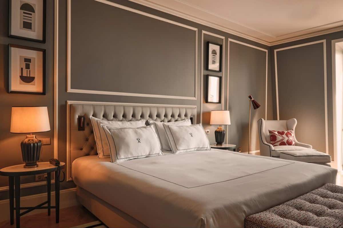 A large bed with a tufted headboard, two bedside tables with lamps, a chair with a cushion, and framed pictures on the wall.