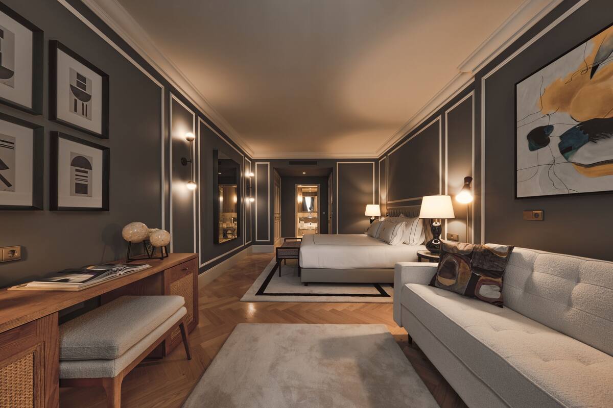 A modern hotel room with a large bed, a sofa, a desk, and a reading area.