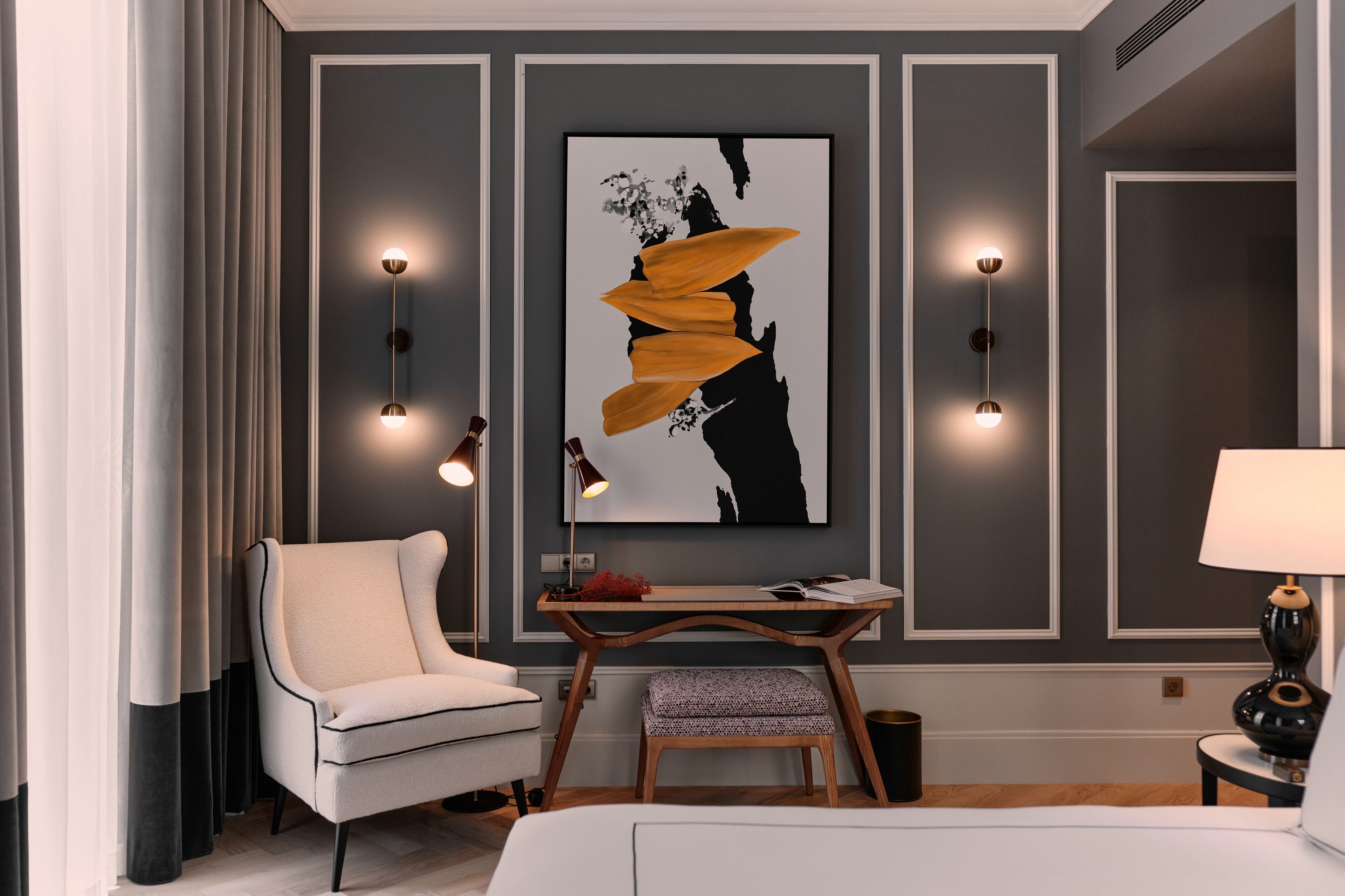 A modern hotel room with a large bed, a white armchair, a small table with a lamp, and a large abstract painting on the wall.