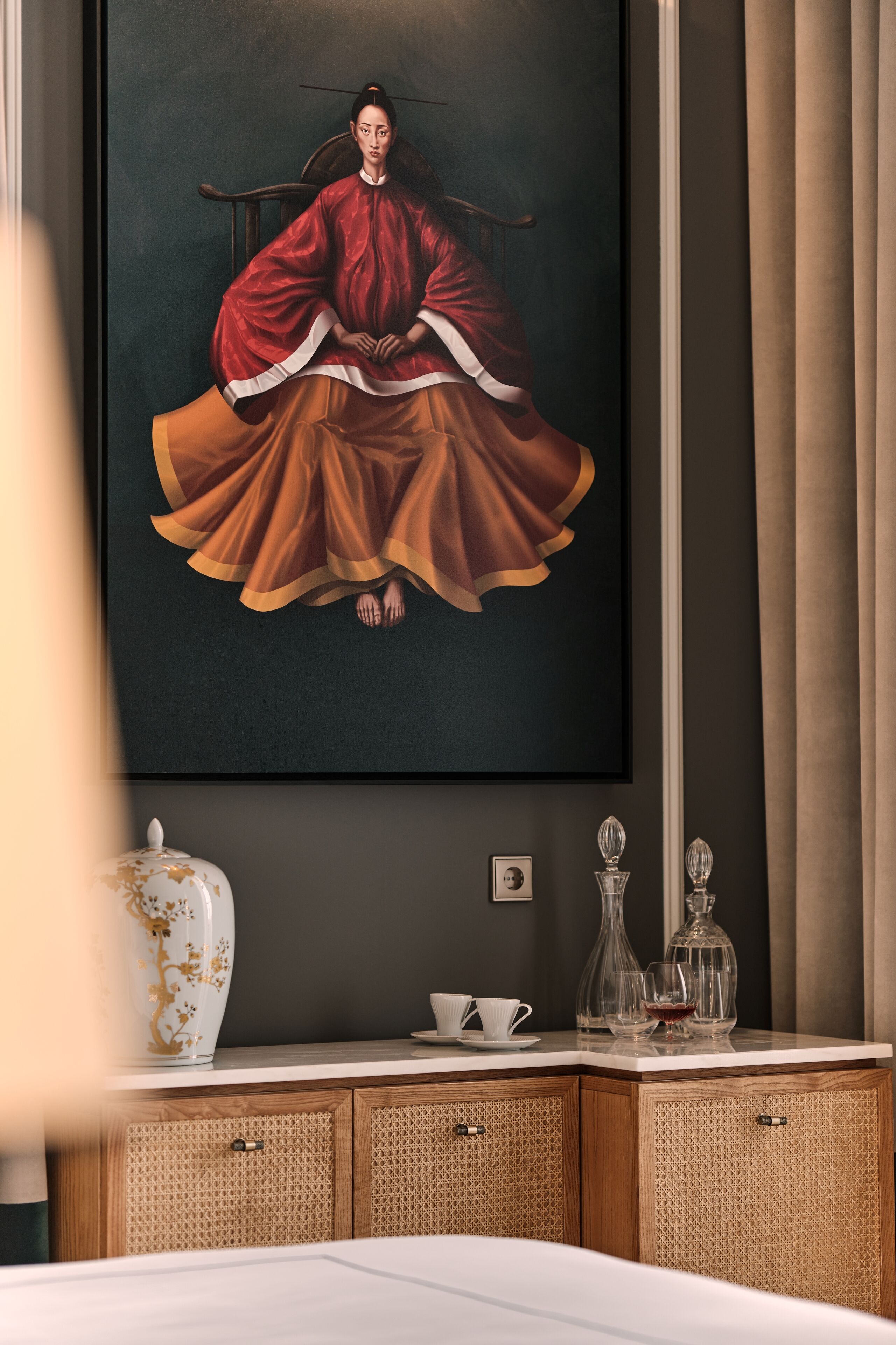 Un tableau encadré représentant une figure vêtue d’un costume traditionnel, un vase blanc orné de motifs dorés et un meuble en bois aux portes tressées.