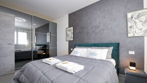 A modern bedroom with a grey quilted bedspread, a teal headboard, and white pillows with text. There is a bedside table with a lamp and a framed artwork on the wall. - Opalia Residence - Eva's Elegance (Peschiera del Garda)