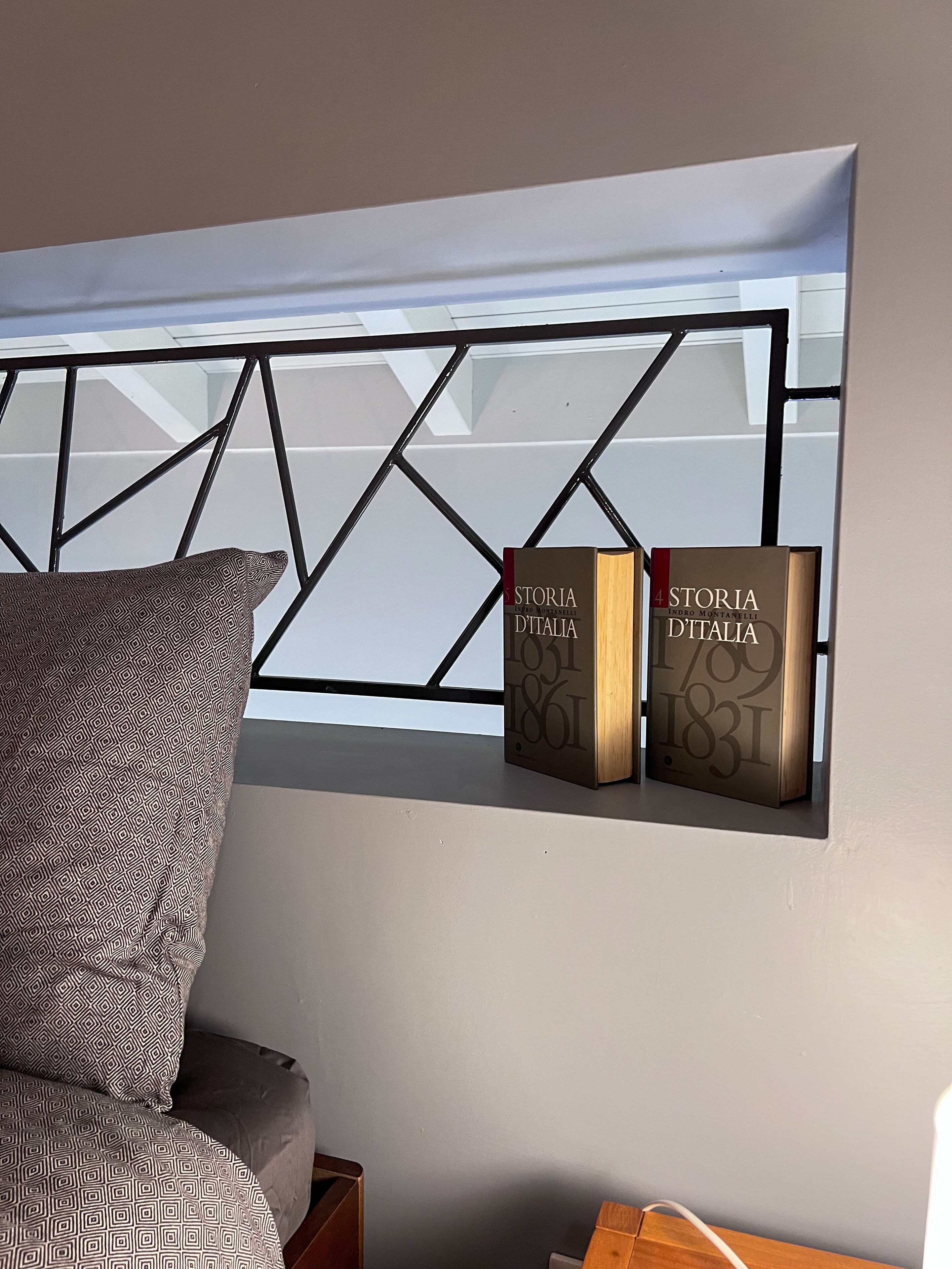 Una habitación moderna con un cojín de estampado geométrico sobre un sofá, una ventana con marco de metal y dos libros titulados 'Storia D'Italia' en una estantería.