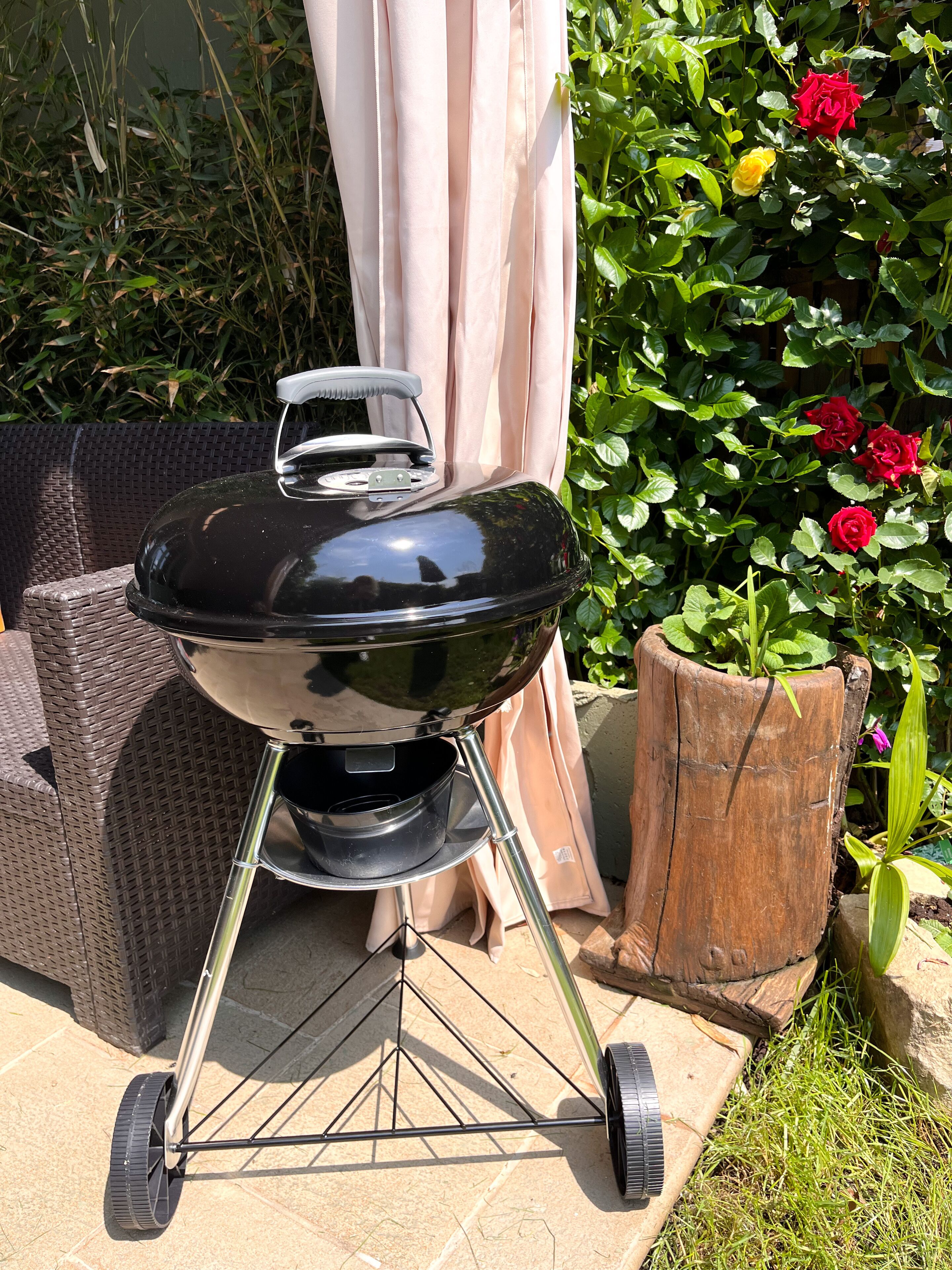 A portable grill with wheels, set on a patio with a wicker sofa and a wooden stump.
