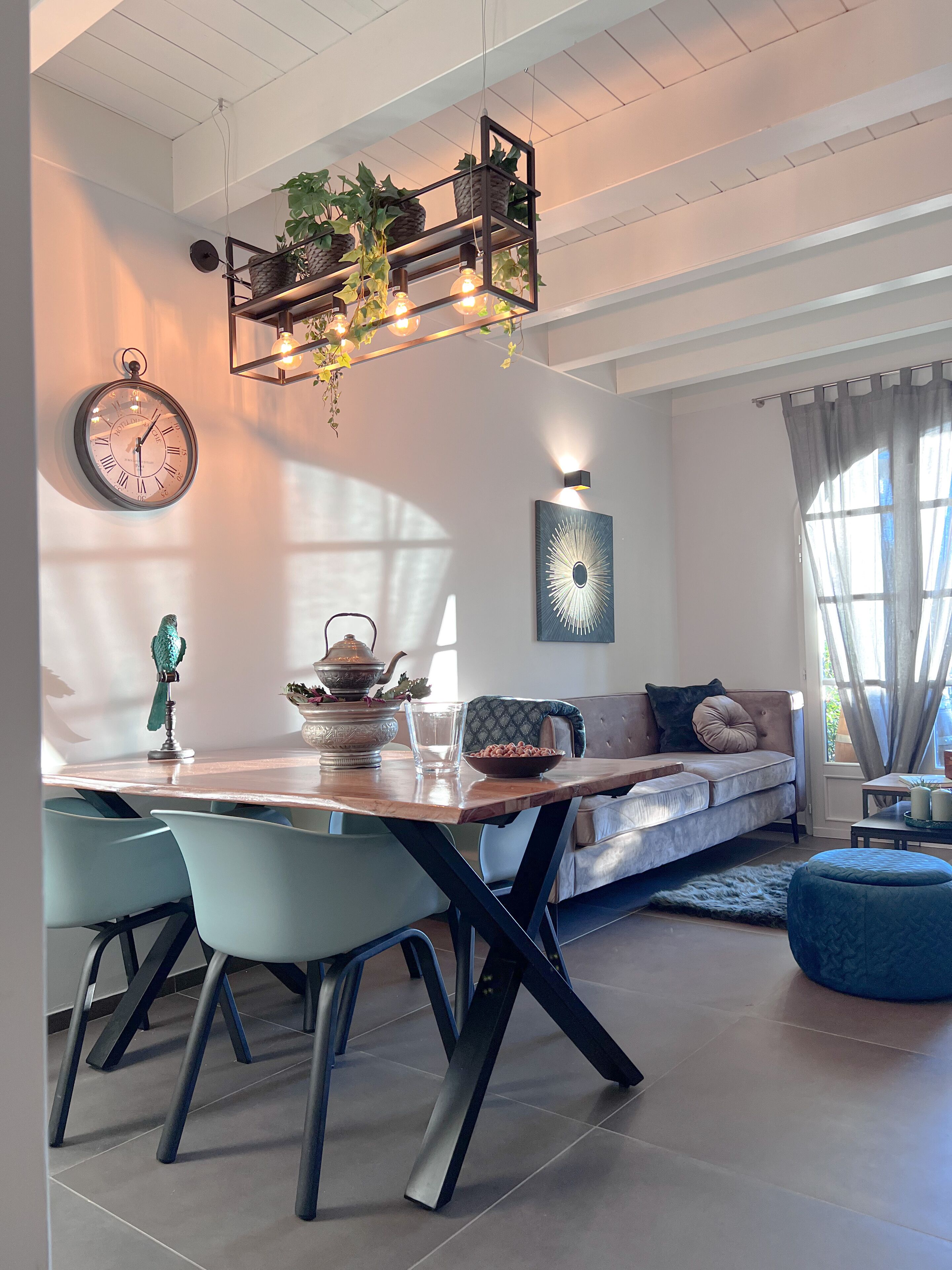 Ein modernes Wohnzimmer mit Essbereich, ausgestattet mit einem Holztisch, hellblauen Stühlen, einer Couch mit Kissen und einem blauen Hocker.
