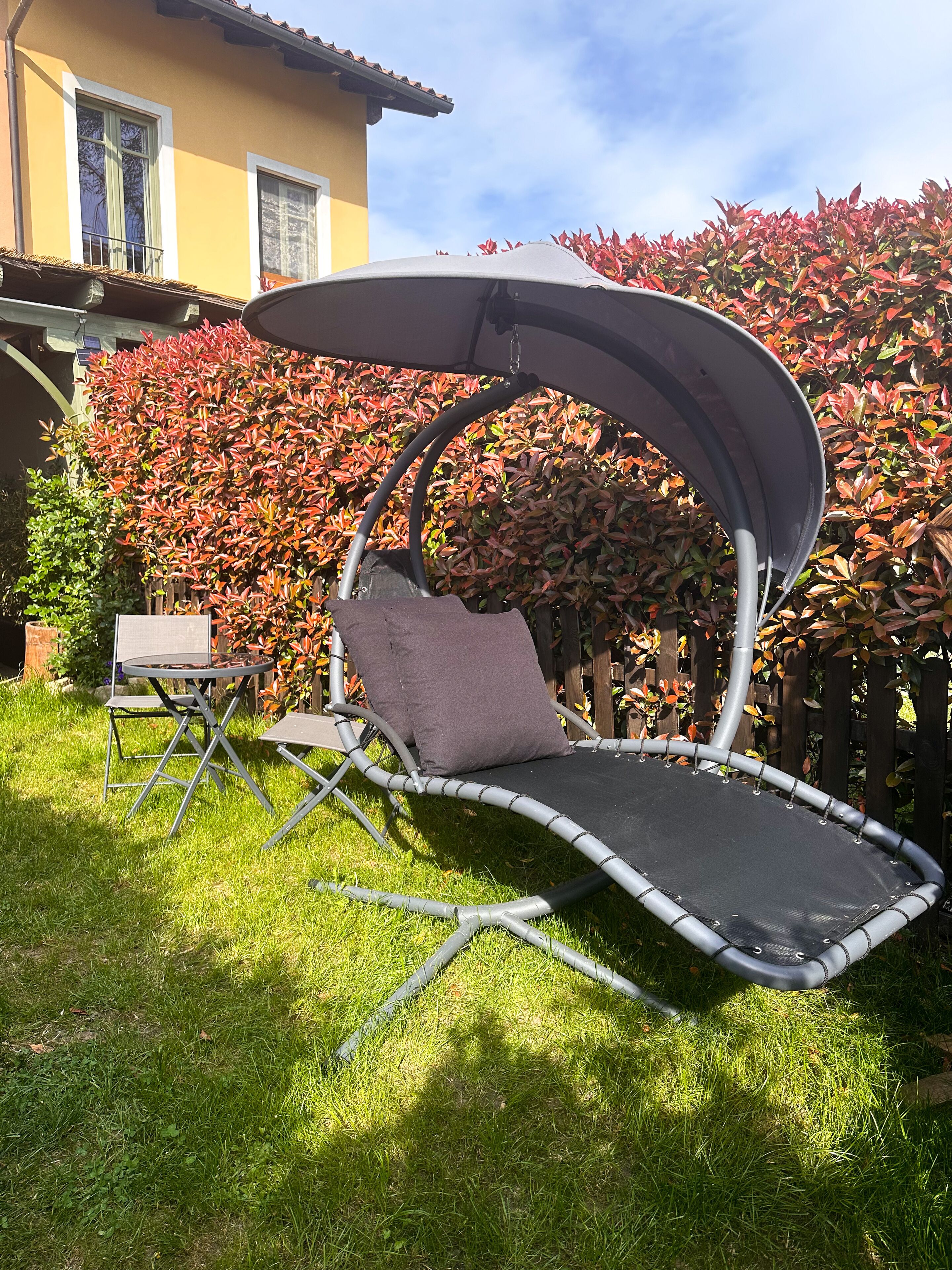 Un trampoline doté d’un auvent, une petite table et une chaise, le tout installé dans une cour verdoyante, avec une maison et des arbustes aux feuilles rouges en arrière-plan.