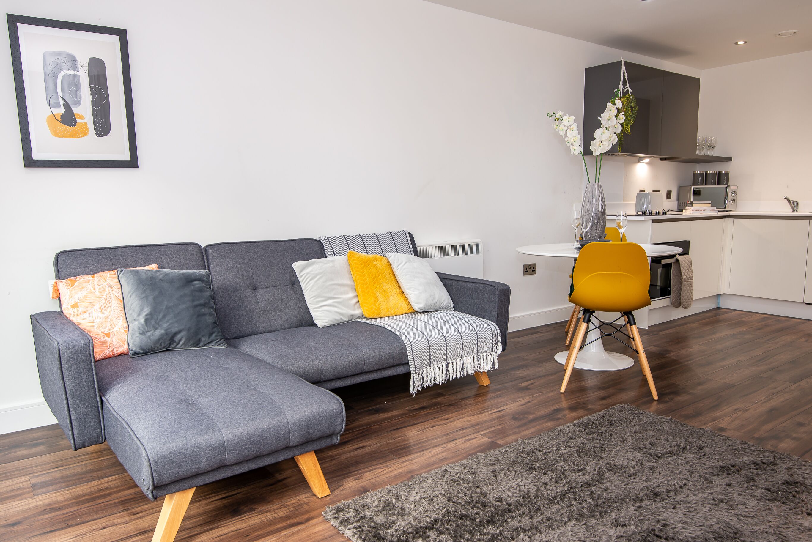 Ein modernes Wohnzimmer mit einem grauen Ecksofa, einem kleinen runden Tisch und einem gelben Sessel. Im Hintergrund ist eine Küche mit einer hängenden Pflanze zu sehen.