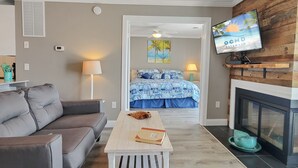 Ett modernt vardagsrum med en grå soffa, ett vitt soffbord med böcker, en säng med ett blått och vitt mönstrat överkast, en tv som är monterad på en vägg av trä och en öppen spis.