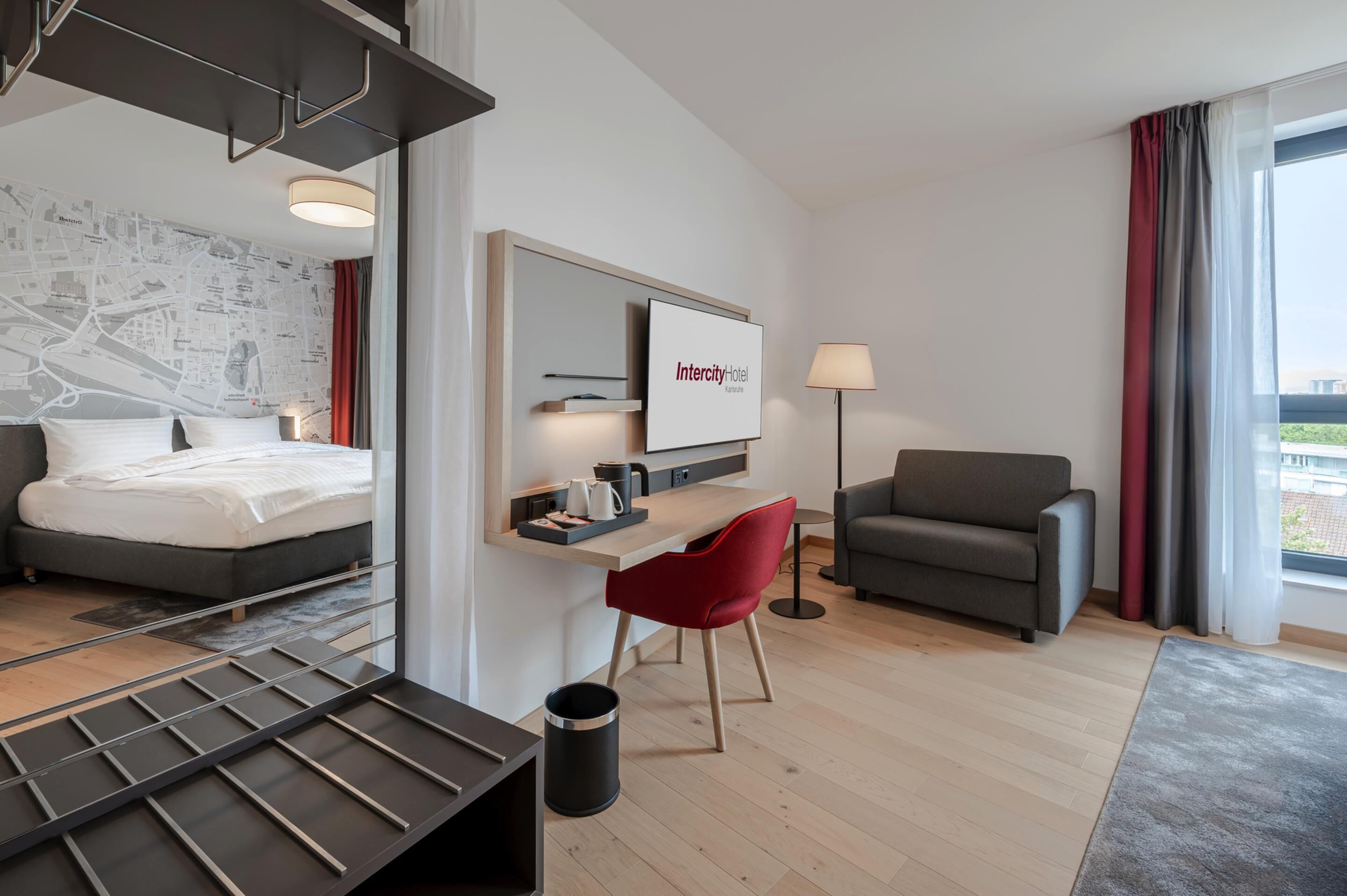 A modern hotel room with a bed, a desk, a red chair, a sofa, and a large window with curtains.