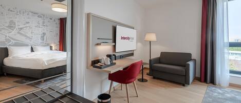 A modern hotel room with a bed, a desk, a red chair, a sofa, and a large window with curtains.
