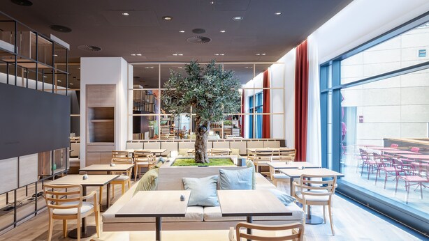 A modern dining area with tables and chairs, a central seating area, and a large window with a view. - IntercityHotel Karlsruhe (Karlsruhe)