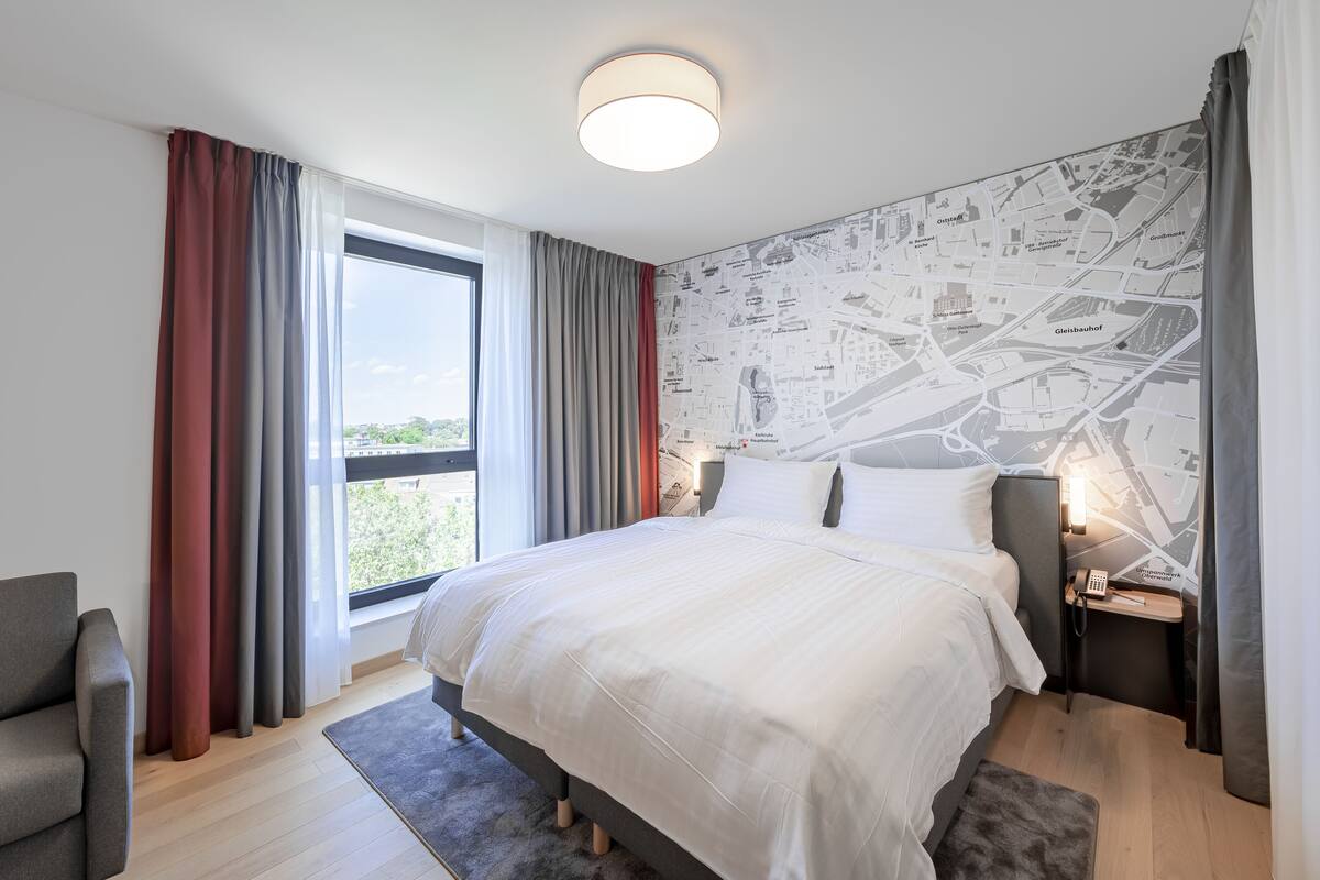 A modern hotel room with a large bed, a map wallpaper, a desk with a lamp, and a view of the city through the window.