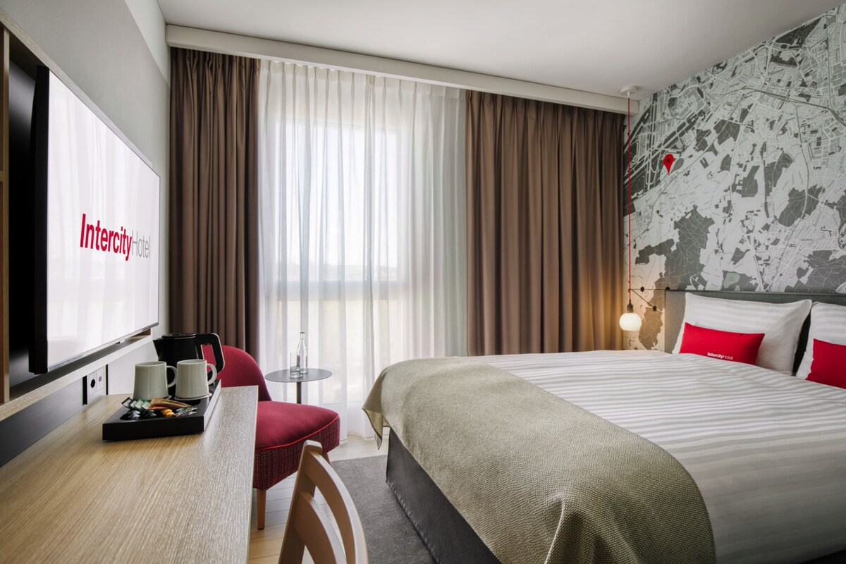 A hotel room with a bed, a desk with a coffee maker, a chair, a TV, and curtains.
