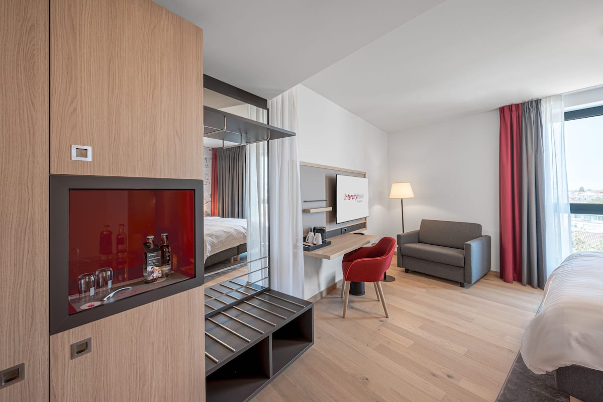 A modern hotel room with a bunk bed, a desk, a red chair, a sofa, a TV, and a window with curtains.