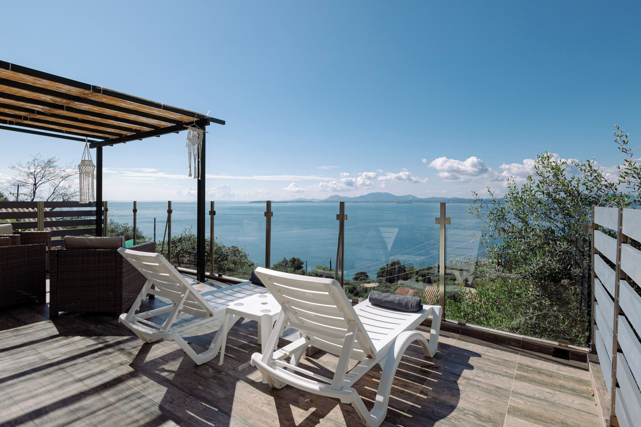 A terrace with two white lounge chairs and a table, overlooking a scenic view of the sea and mountains.