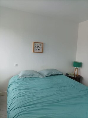 Een slaapkamer met een bed, kussens, een ingelijst schilderij aan de muur, een nachtkastje met een lamp en een boek.