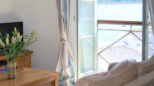 A room with a large window, a wooden table with a vase of flowers, and a bed with a view of the sea. - Crows Nest: a modern apartment in the centre of St Mawes with stunning sea views (St Mawes)