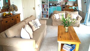 A living room with two beige sofas, a wooden coffee table, and a bookshelf filled with books. - Crows Nest: a modern apartment in the centre of St Mawes with stunning sea views (St Mawes)