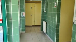 A corridor with green walls, yellow doors, and tiled flooring. There are signs indicating restrooms and a fire exit. - Electrified Camping Pitch (Bessé-sur-Braye)