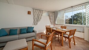 Een kamer met een grijze bank, een houten eettafel en stoelen. Er is een raam met een lattenroede en gordijnen.