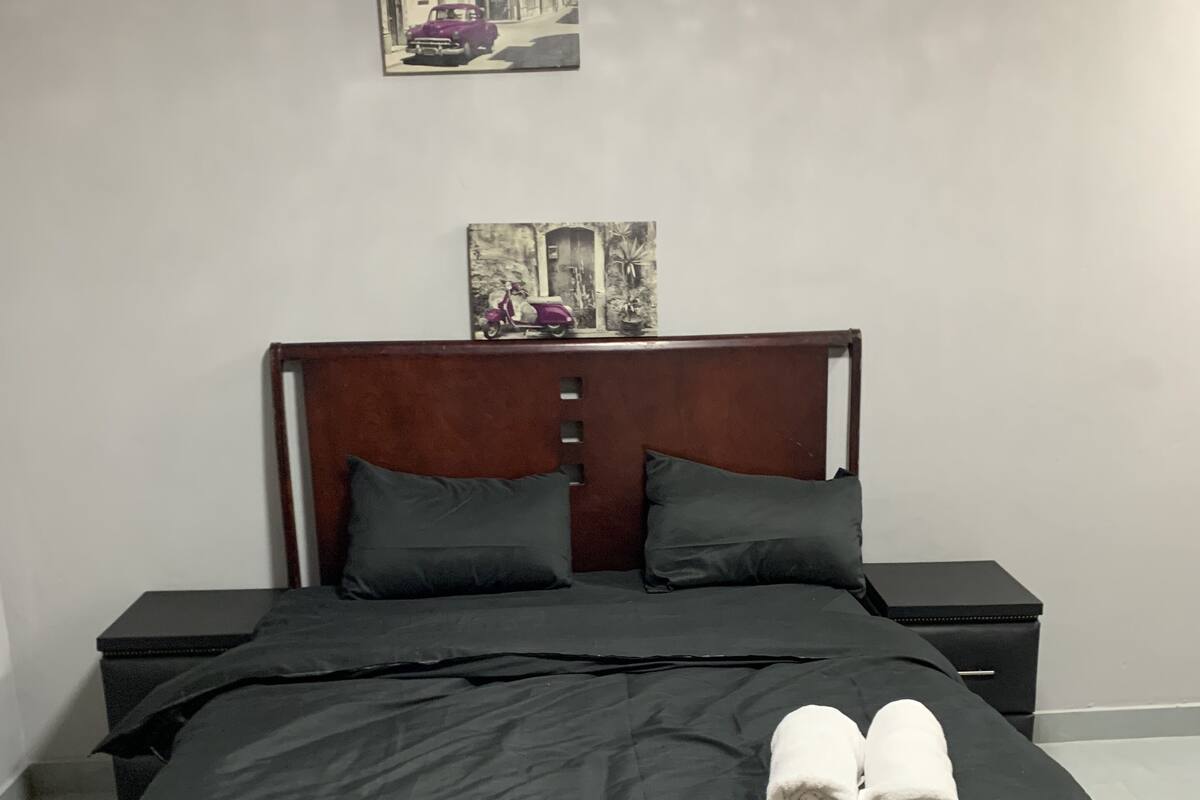 Ein ordentlich bezogenes Bett mit dunkler Bettdecke und Kissen, zwei Nachttische mit schwarzer Oberfläche sowie ein an der Wand hängendes Bild über dem Bett.