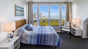 A hotel room with a large bed, a chair, a nightstand, and a view of the beach. - Vacation Rental in the Bayfront Community of Port Lewes (Lewes Beach)