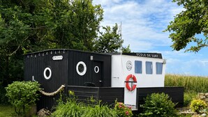 A floating structure with a black and white exterior, labeled 'ECOLE DE SURF,' surrounded by lush greenery and a clear sky. - Authentic Fisherman’s Cabin • Audierne Bay • Brittany • 2 Guests (Saint-Jean-Trolimon)
