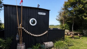 A black wooden structure with a circular window, a flag, and a sign reading 'GV.2017'. - Authentic Fisherman’s Cabin • Audierne Bay • Brittany • 2 Guests (Saint-Jean-Trolimon)