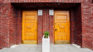 Dos puertas de madera con esculturas de león, una maceta blanca con planta y un muro de ladrillo.