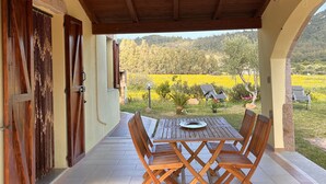 A covered patio with a wooden table and chairs, a view of a garden with yellow flowers, and a stone wall. - Villa Patrizia (Castiadas)