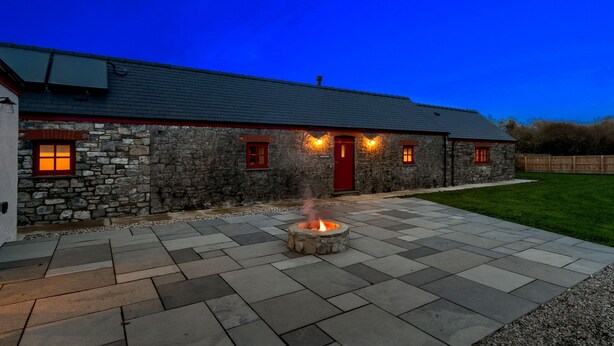 A stone building with a red door and windows, a fire pit in the center, and a paved patio area. - Stylish barn nestled in a scenic setting (Laugharne)