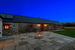 A stone building with a red door and windows, a fire pit in the center, and a paved patio area. - Stylish barn nestled in a scenic setting (Laugharne)