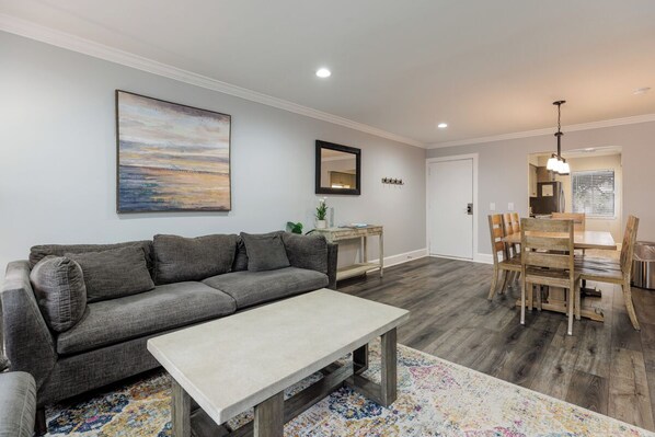 Una sala de estar moderna con un sofá gris, una mesa de comedor de madera con sillas y un cuadro grande en la pared.