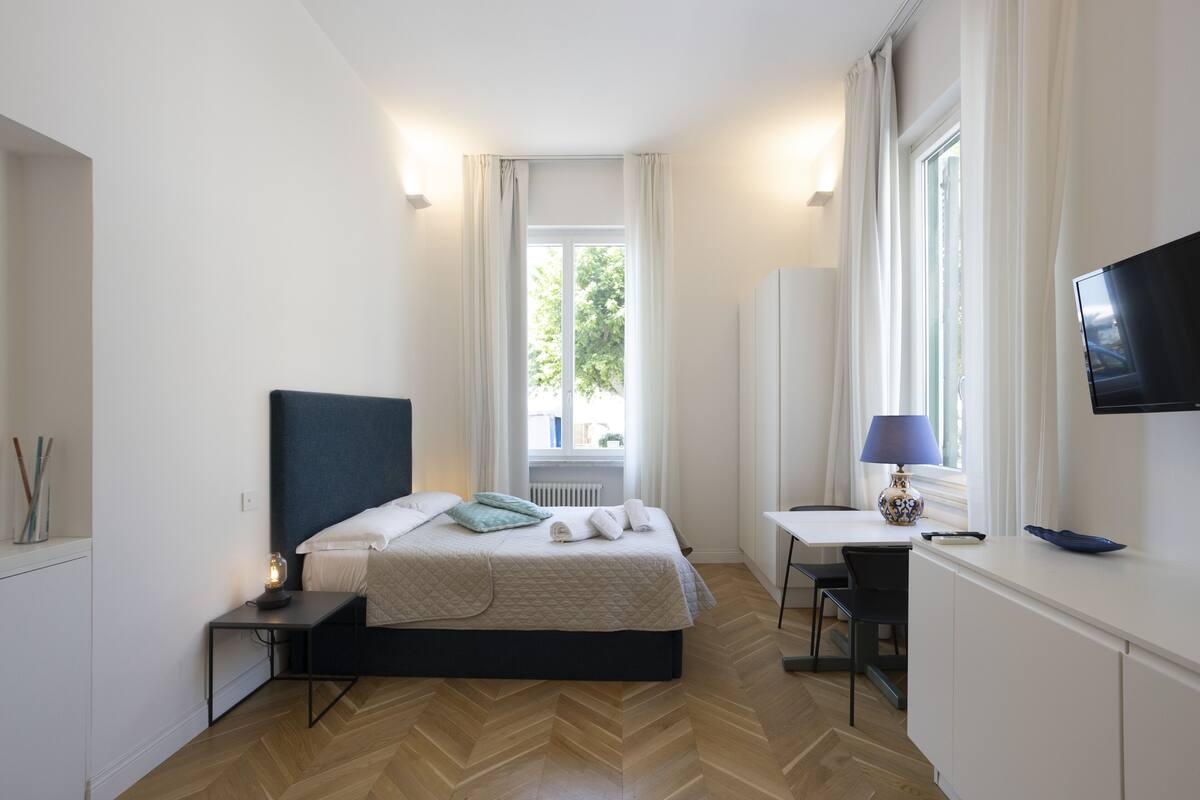 A modern hotel room with a bed, bedside table, lamp, TV, and a view of greenery outside.