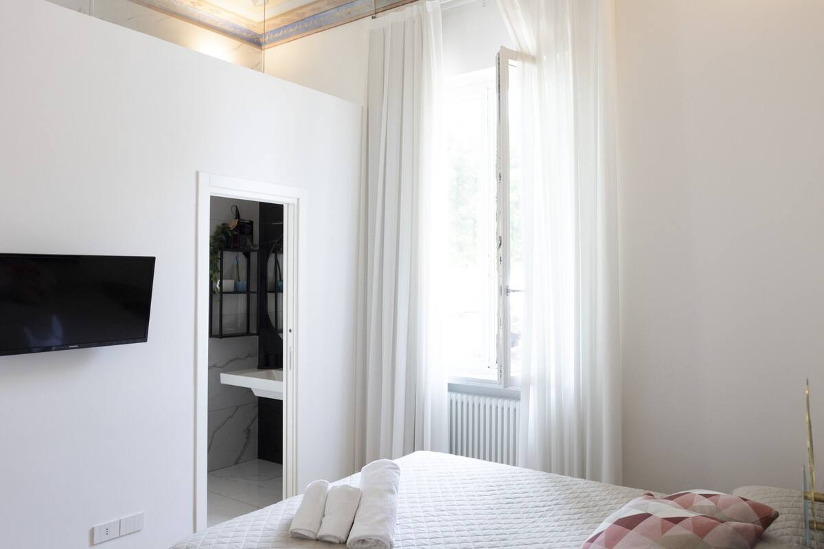 A bedroom with a bed, a TV mounted on the wall, a window with curtains, and a ceiling with a floral pattern.