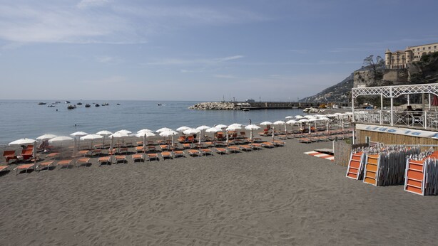 A beach with rows of sunbeds and umbrellas, a pier, and a castle-like building in the background. - Sinonda Luxury Apartment (Maiori)