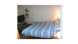 Ein Schlafzimmer mit einem Bett, einem Fernseher auf einem Schrank, einer Lampe und einem kleinen Tisch mit einer Fernbedienung.
