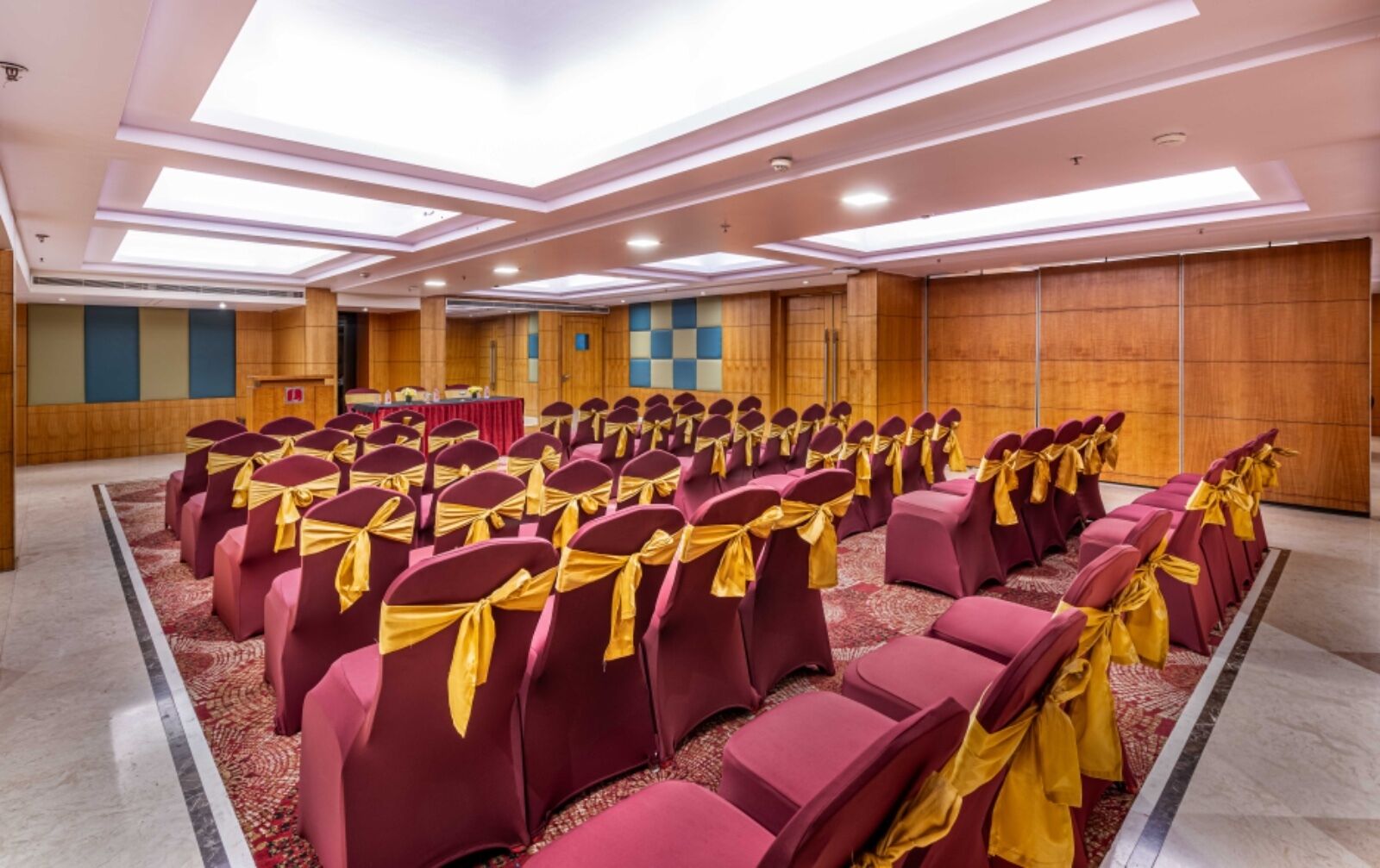 A conference room with rows of chairs, each with a gold sash, and a stage with a red table and chairs.