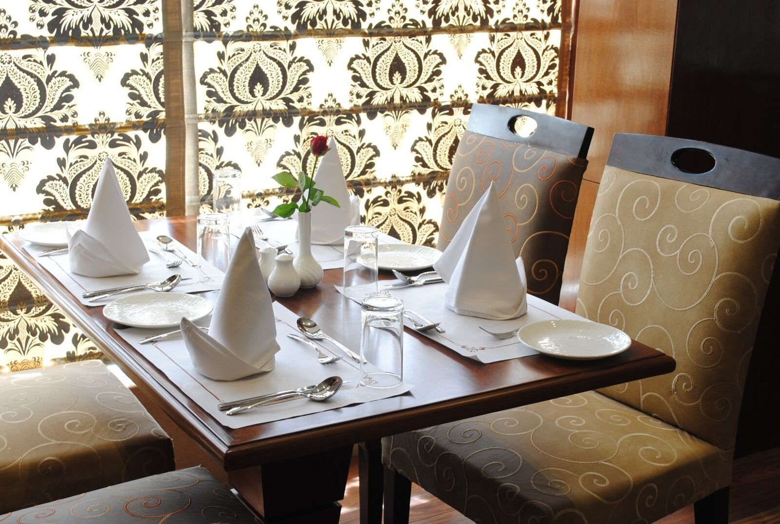 Una mesa de comedor dispuesta con servilletas blancas, platos y cubiertos, con un jarrón de flores y un vaso de agua.