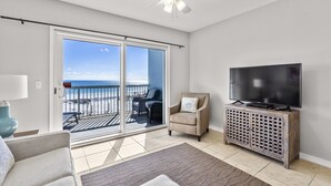 Ein Wohnzimmer mit Fernseher, Sessel, Sofa, Deckenventilator und Blick auf den Ozean durch eine Schiebetür.