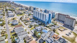 Vue aérienne d’une zone hôtelière côtière comprenant plusieurs bâtiments, dont un hôtel imposant à la façade bleue et blanche, situé près d’une plage.