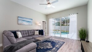 Ein Wohnzimmer mit grauem Ecksofa, einem Holztisch, einem blauen Teppich mit Muster, einem Deckenventilator und Blick auf einen Swimmingpool durch große Glasschübe.