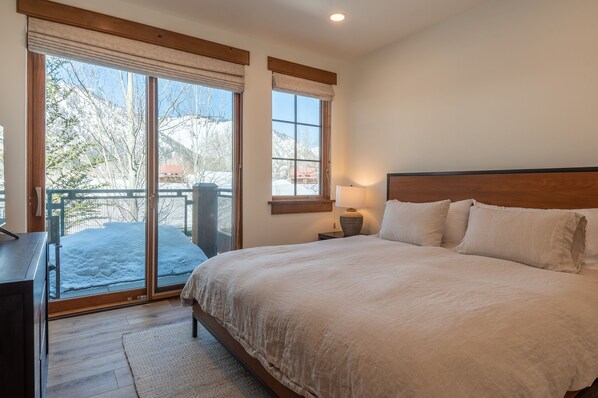 A bedroom with a large bed, a nightstand, a lamp, and a view of a snowy outdoor area through a sliding glass door. - Westridge Condo 26 -Updated Condo w/ AC, Walk to River Run Lifts and Downtown (Ketchum)