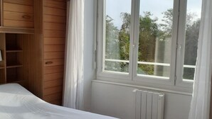 A bedroom with a bed, wooden wardrobe, a window with curtains, and a radiator. - Agréable Appartement Dans Résidence Avec Jardin Très Proche des Commodités (Pont-l'Abbé)