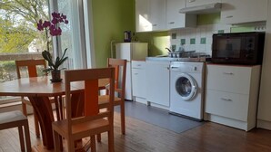 A compact kitchen with a washing machine, a microwave, and a dining area with wooden chairs and a table. - Agréable Appartement Dans Résidence Avec Jardin Très Proche des Commodités (Pont-l'Abbé)