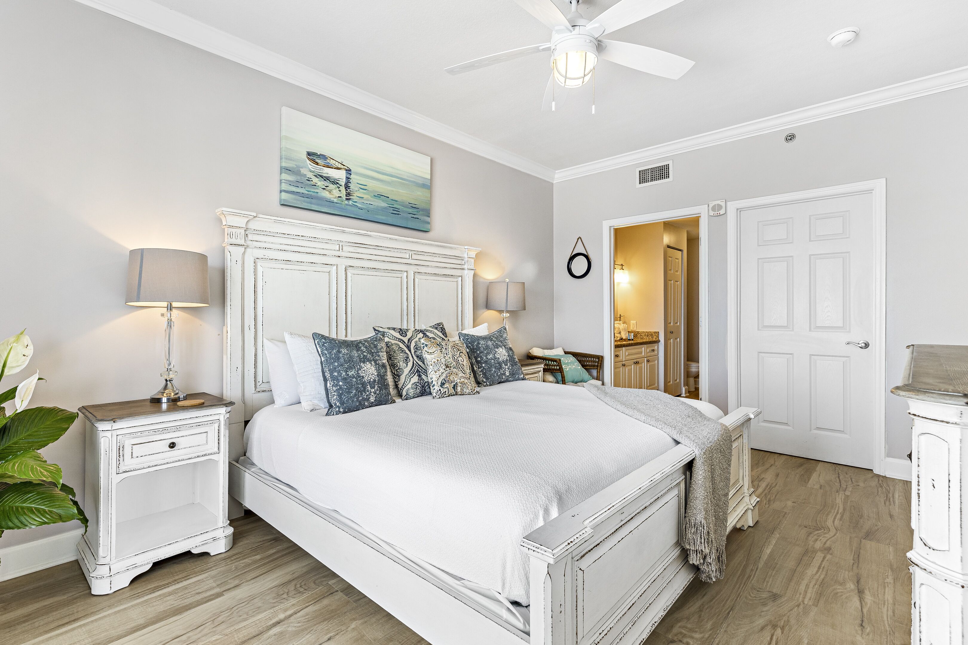 A bedroom with a white bed, a nightstand with a lamp, a ceiling fan, and a painting above the bed.