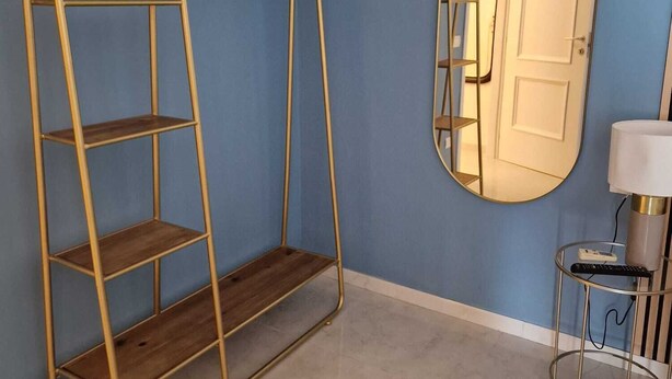 A room with a blue wall, a gold metal shelf unit, a round mirror, and a small table with a lamp. - Art Flat Municipio (Naples)